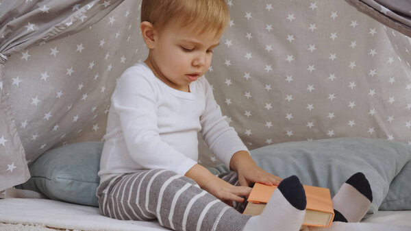 curious toddler boy sitting in baby wigwam and holding book