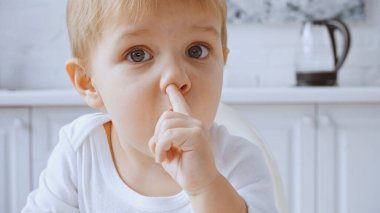 close up of toddler boy picking nose while looking at camera at home