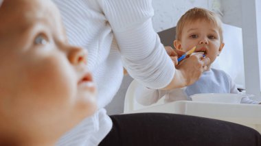 mother feeding distracted toddler boy looking at sister on blurred foreground 