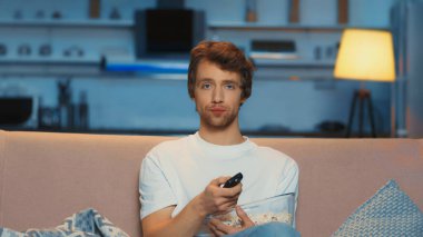 young man holding remote controller while clicking channels and watching tv on couch
