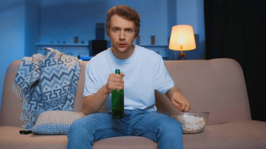 young man with bottle of beer sitting on couch near bowl of popcorn while watching championship  