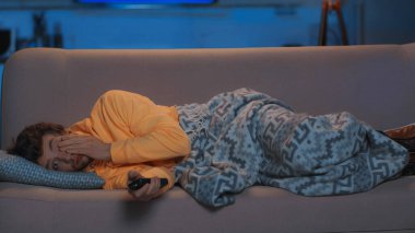sleepy man in yellow hoodie lying under blanket and watching tv on sofa at night