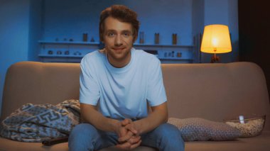 cheerful young man in white t-shirt sitting on sofa near bowl with popcorn in living room