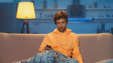skeptical young man in yellow hoodie holding remote controller and clicking channels 