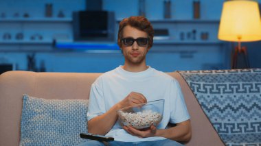 young man in 3d glasses holding bowl with popcorn while watching movie in living room 