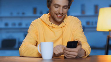 cheerful young man messaging on smartphone near cup of tea on wooden table 