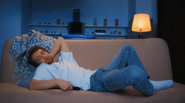 sleepy young man lying on couch with remote controller in modern living room at night  clipart