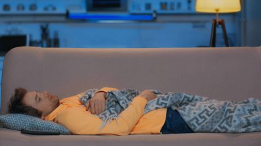 young man in yellow hoodie sleeping under blanket near remote controller at night clipart