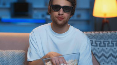 young man in 3d glasses holding bowl with popcorn while watching film in living room