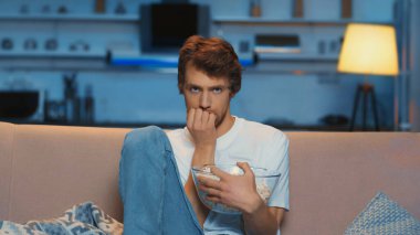 tensed young man holding bowl of popcorn while watching scary movie 