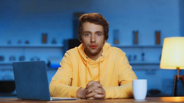young man in yellow hoodie sitting with clenched hands near laptop and cup of coffee in evening 