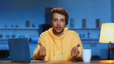 young man in yellow hoodie talking while gesturing near laptop and cup of coffee in evening 