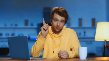 young man in yellow hoodie pointing with finger near laptop and cup of coffee in evening 