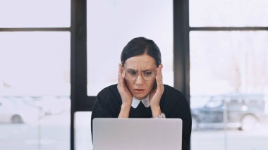 worried businesswoman in eyeglasses touching head and looking at laptop in office