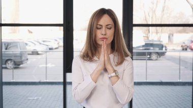 tense woman with closed eyes standing with praying hands before job interview