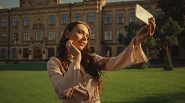 Brunette woman taking selfie on smartphone in summer park during sunset