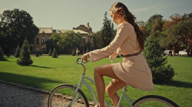 Side view of brunette woman in dress cycling in summer park 