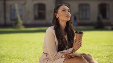 Dreamy young woman holding paper cup and book in summer park 