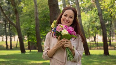 Smiling woman holding bouquet and looking at camera in park 