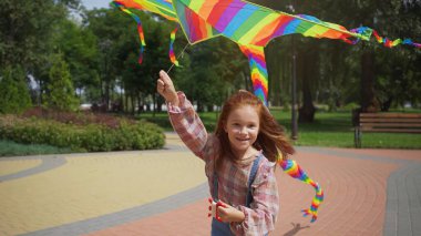 Cheerful redhead girl running with flying kite in park 