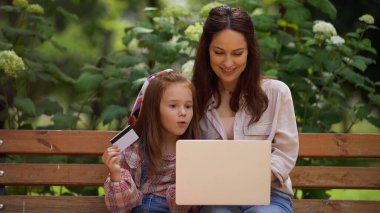 Smiling woman using laptop near near excited daughter with credit card on bench in park 