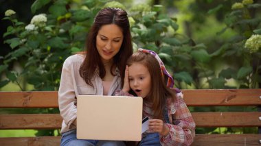 Shocked kid holding credit card near mom using laptop on bench in park 