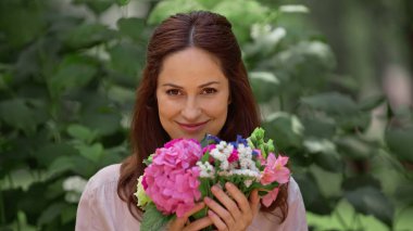 Cheerful brunette woman looking at camera and holding flowers in park 