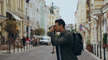 Side view of bi-racial traveler taking photo on film camera on european street 