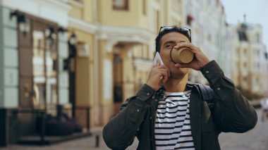 Bi-racial man talking on smartphone and drinking coffee to go on urban street 