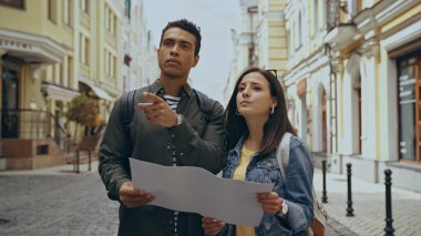 Bi-racial tourist pointing with finger near girlfriend with map on urban street 