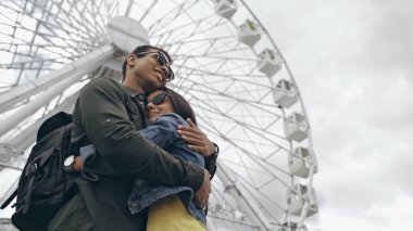 Low angle view of bi-racial tourist hugging girlfriend near ferris wheel 