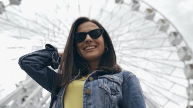 Low angle view of smiling tourist in sunglasses standing near blurred ferris wheel 