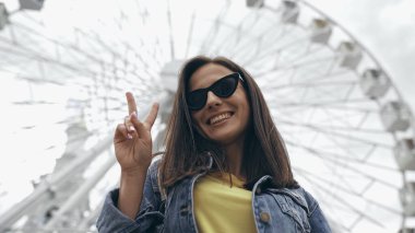 Low angle view of cheerful traveler showing peace sign near blurred ferris wheel 