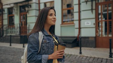 Positive tourist with backpack holding coffee to go on urban european street 
