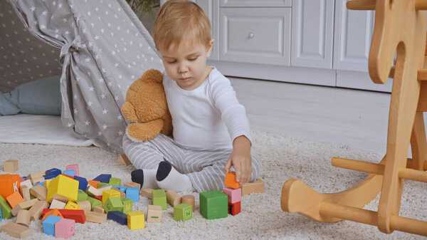 toddler boy holding teddy bear and playing with multicolored wooden blocks near rocking horse on carpet 