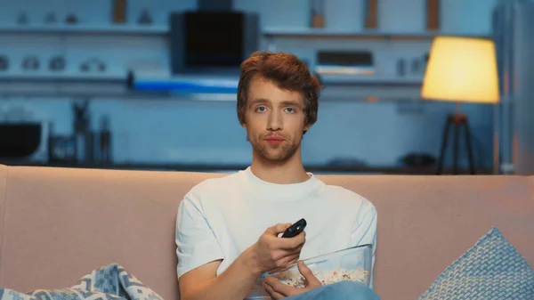 young man holding remote controller while clicking channels and watching tv on couch