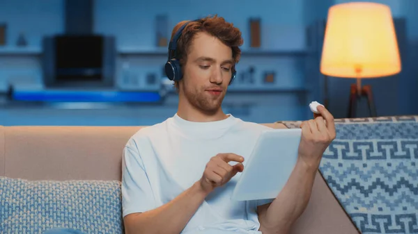 young man in wireless headphones using digital tablet and holding marshmallow in living room