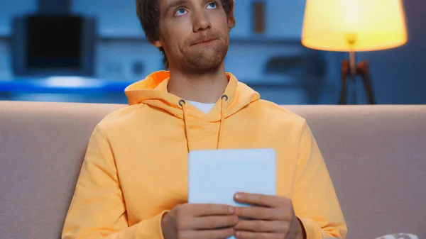 pensive young man in yellow hoodie holding digital tablet in living room