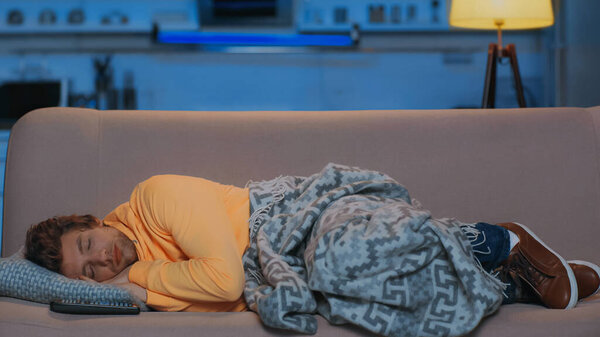 young man in yellow hoodie sleeping under blanket near remote controller at night 