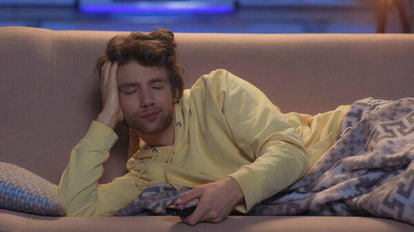 tired young man with closed eyes resting under blanket and holding remote controller at night