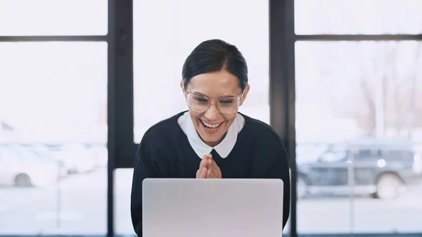 joyful businesswoman in eyeglasses showing wow gesture with folded hands near laptop