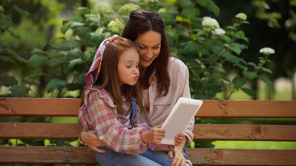 Child pouting lips near smiling mom while using digital tablet on bench in park 