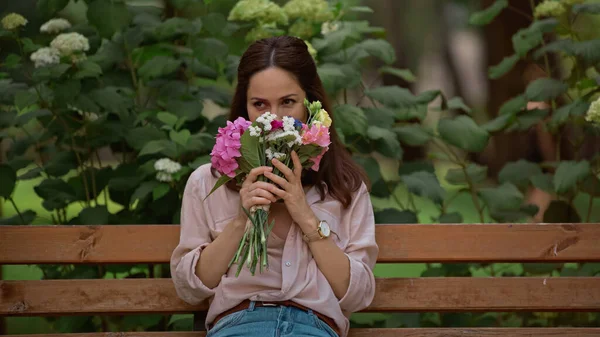 Brunette woman smelling bouquet while sitting on bench in park 