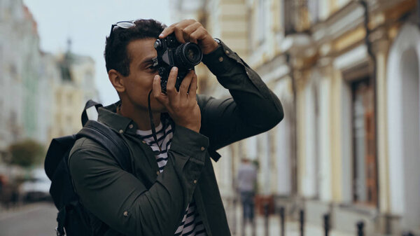 Bi-racial traveler with backpack taking photo on vintage camera on urban street 