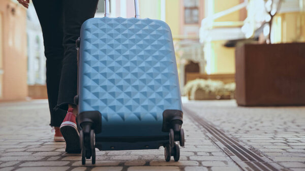 Cropped view of traveler walking near suitcase on urban street 