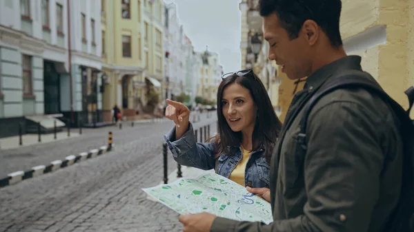 Smiling tourist pointing with finger near bi-racial boyfriend with map on urban street