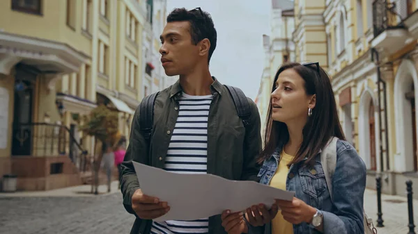 Young interracial couple holding map and looking away on urban street