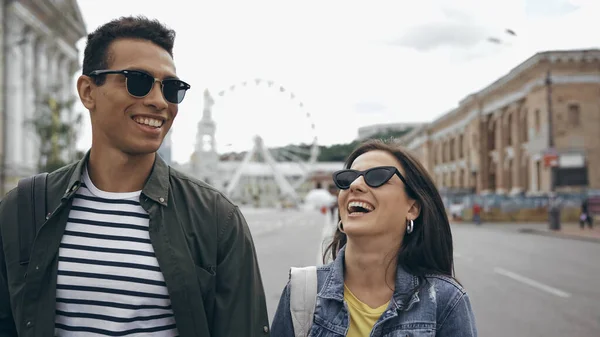 Positive interracial tourists in sunglasses walking on urban street