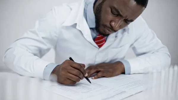 Focused african american doctor in white coat signing documents in clinic — Stock Photo