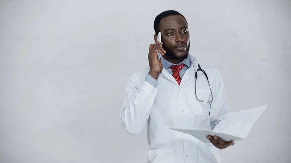 Afrikanisch-amerikanischer Arzt spricht auf Smartphone und hält Ordner mit Dokumenten in der Hand — Stockfoto